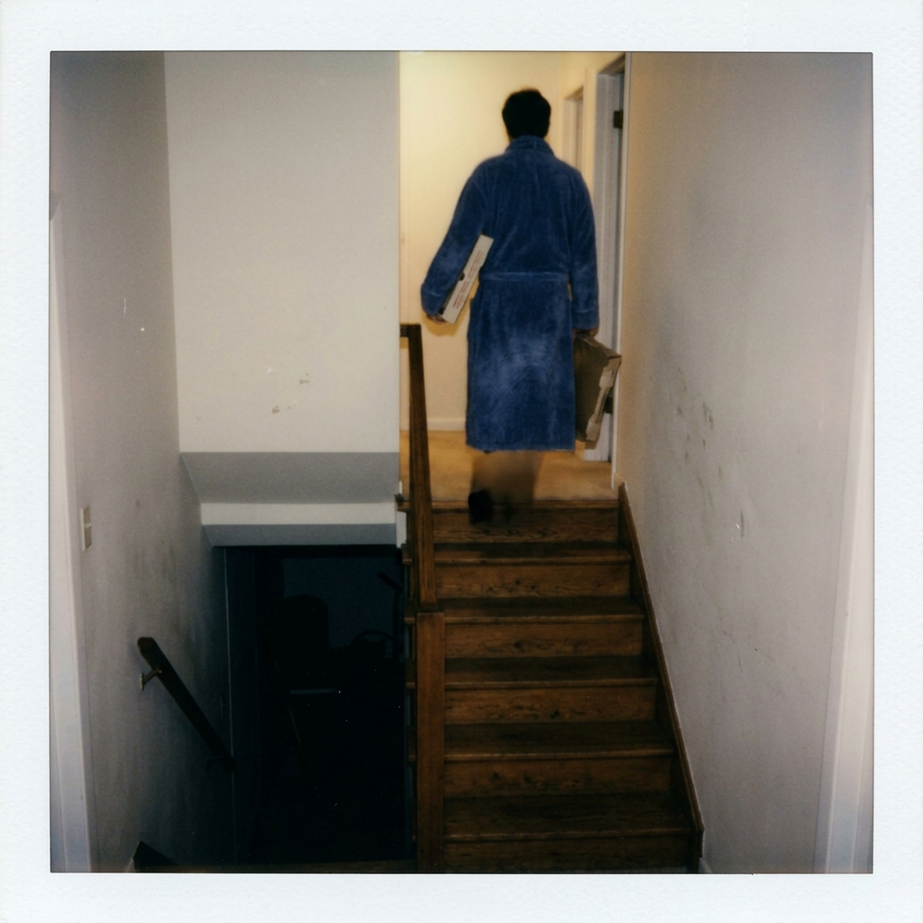 A parent in a robe at the top of basement stairs at night, backlit, holding an empty pizza box